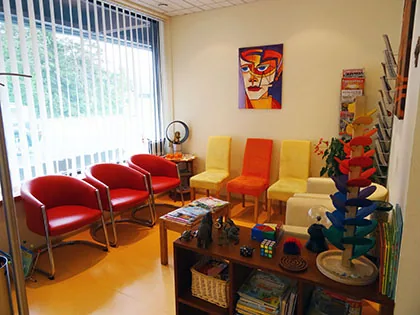 Wartezimmer mit roten, orangen und gelben Stühlen, einem bunten Porträt an der Wand, Kugelbahn und Spielsachen